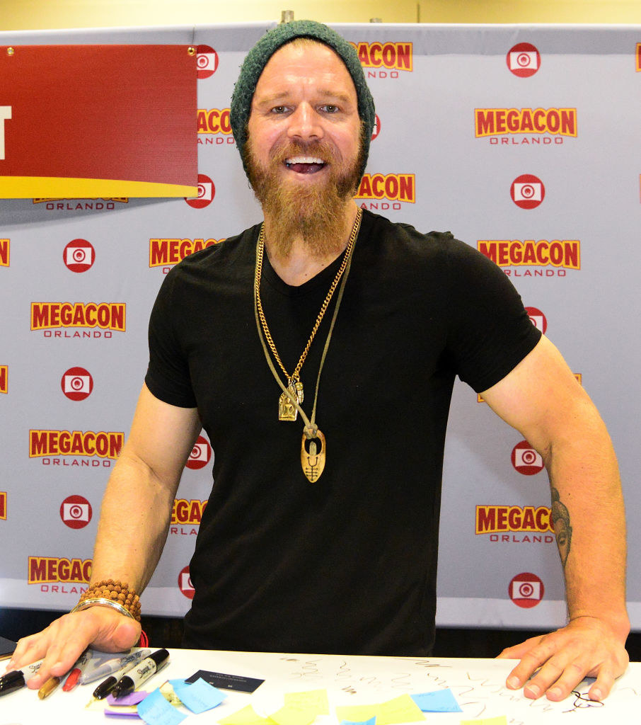 wearing a beanie, t-shirt, and gold necklaces, the actor smiles from his booth at a fan convention
