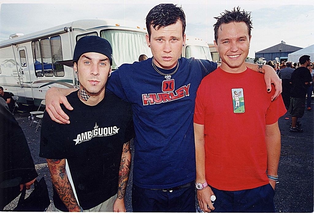 Band members Mark Hoppus, Travis Barker, and Tom DeLonge pose in front of a trailer during a music festival