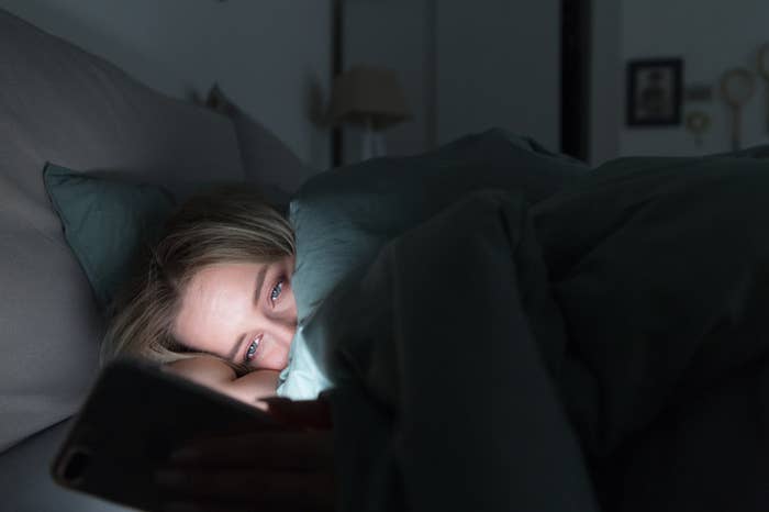 A person in bed under the covers staring at their phone