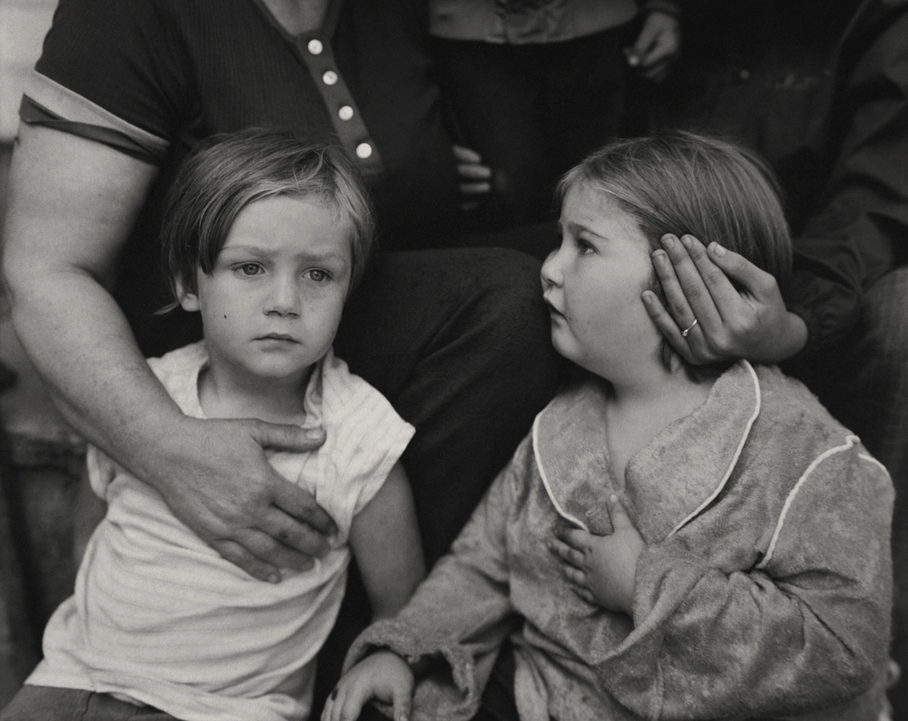 two young girls sit with adults who are cropped out of the frame, whose arms extend down to touch the girls