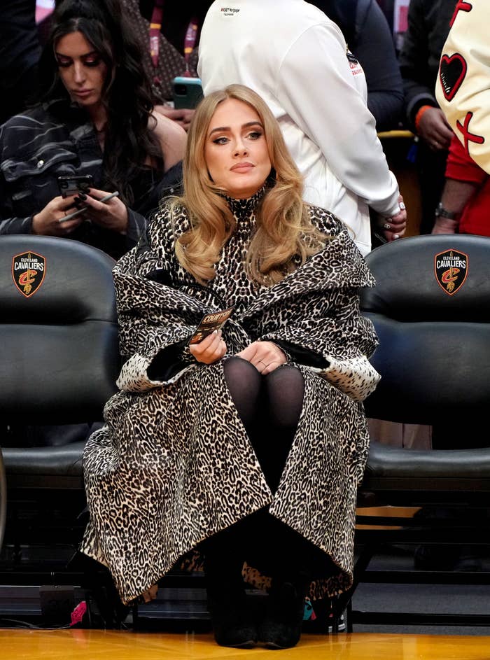 Adele sitting court side in a leopard print jacket and tights