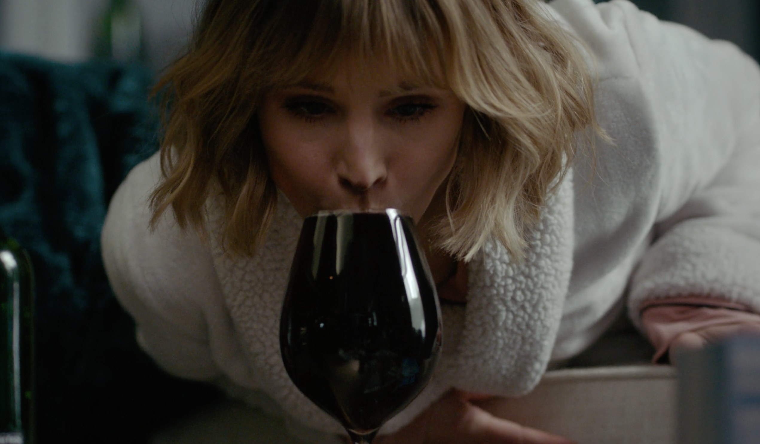 Kristen Bell in a bathrobe leaning down to slurp red wine from a glass filled to the brim