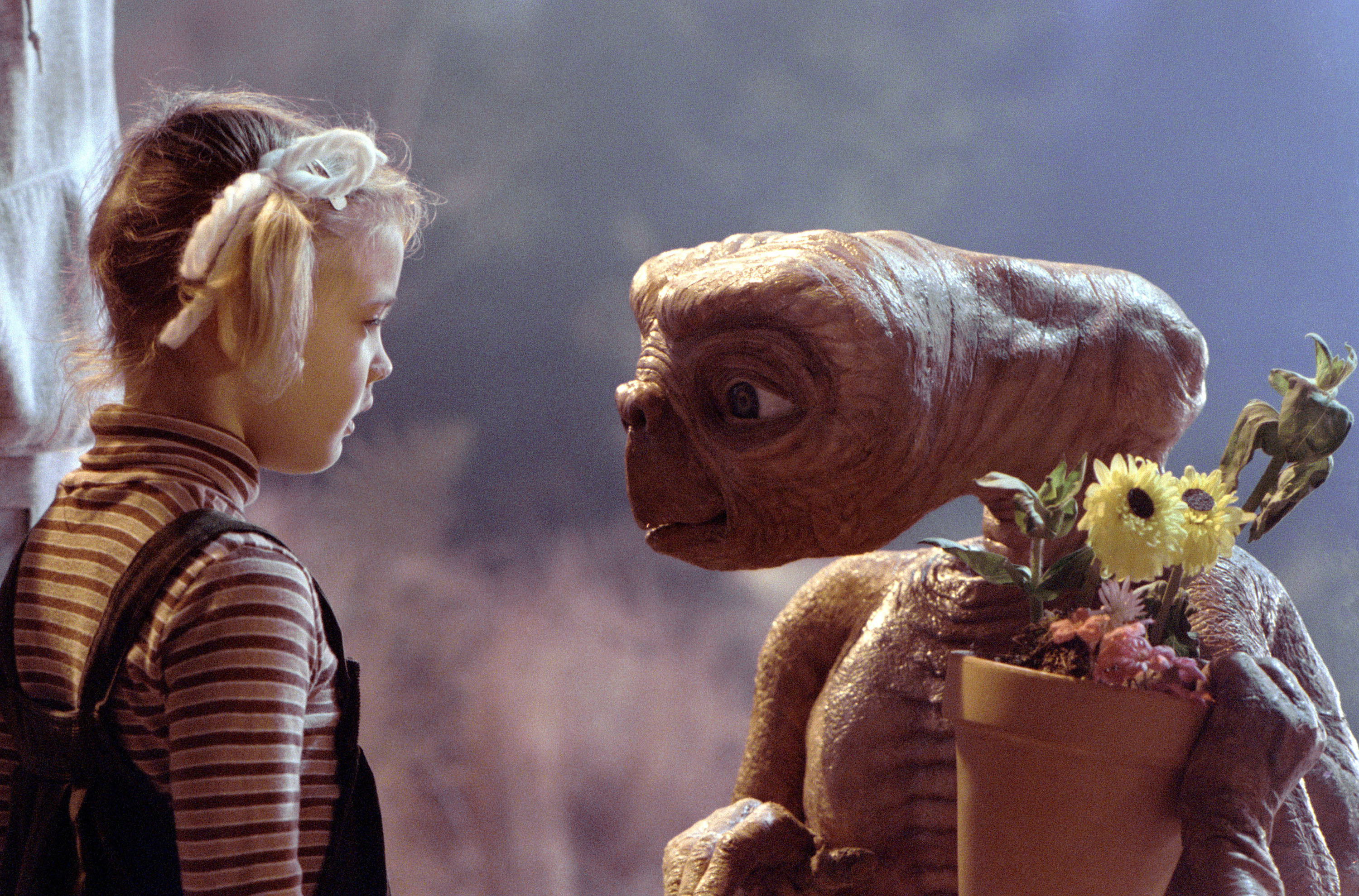 Drew Barrymore stands in front of E.T., who is holding a pot of flowers