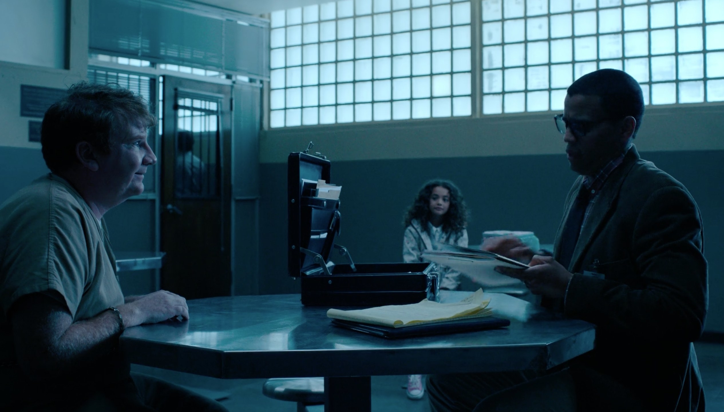 A prisoner (left) across from a psychiatrist (right) and the psychiatrist&#x27;s daughter in the background of an interrogation room