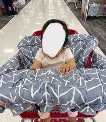 reviewer's photo of their child sitting in the gray shopping cart cover at the store