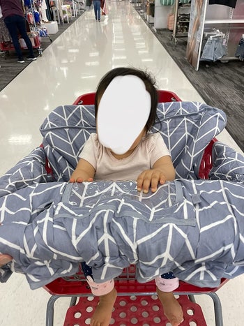 reviewer's photo of their child sitting in the gray shopping cart cover at the store