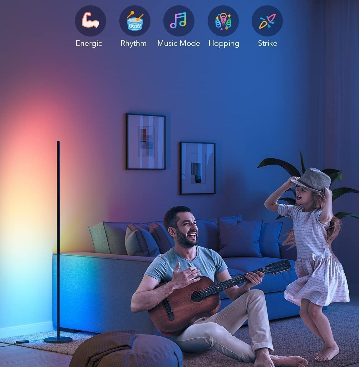 A person playing a guitar in their living room while a child dances to the music with the lamp casting a sunset glow onto the wall