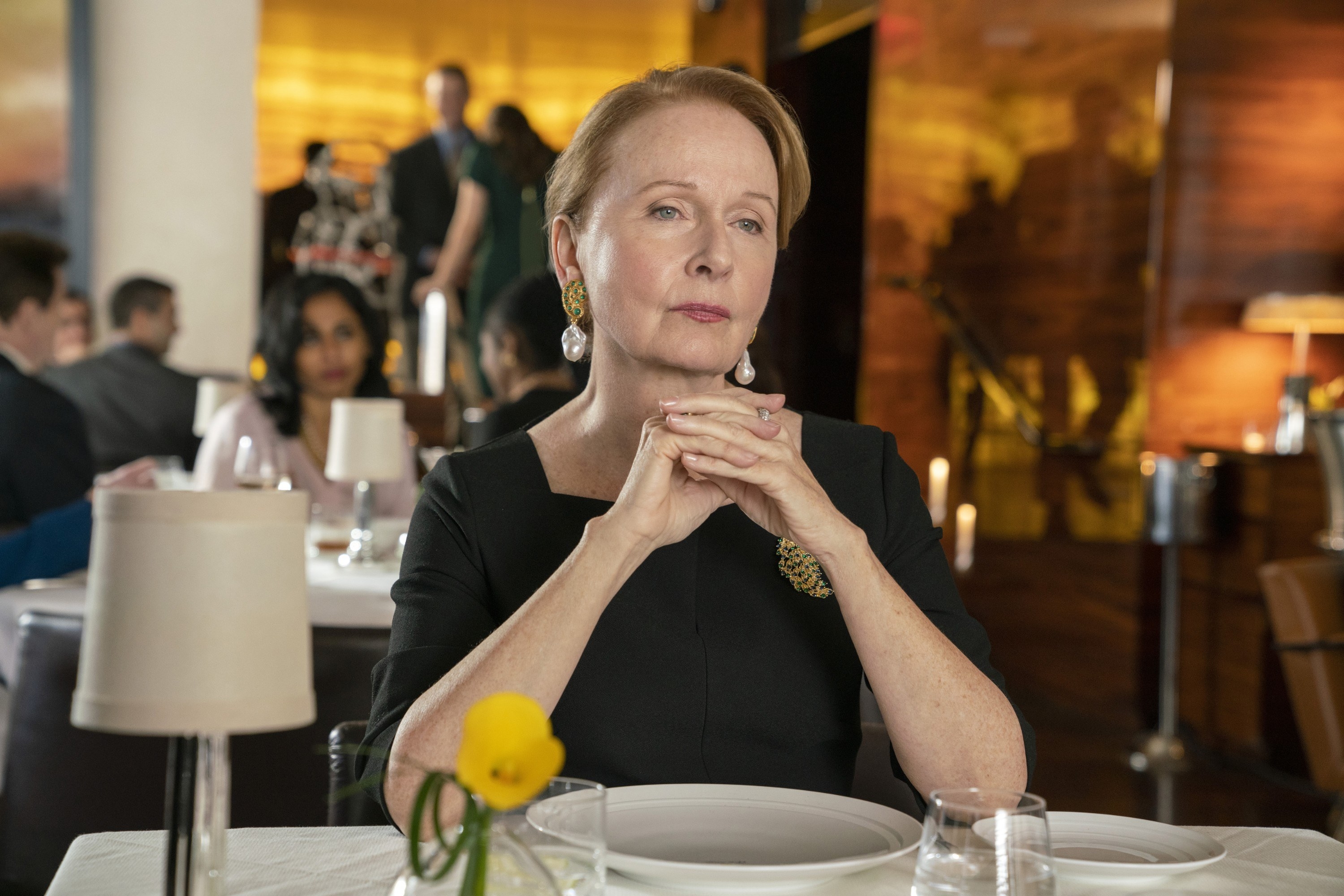 wearing an elegant dress with a square neckline and gaudy earrings and a brooch, Nora sits with her hands folded at a fancy restaurant table