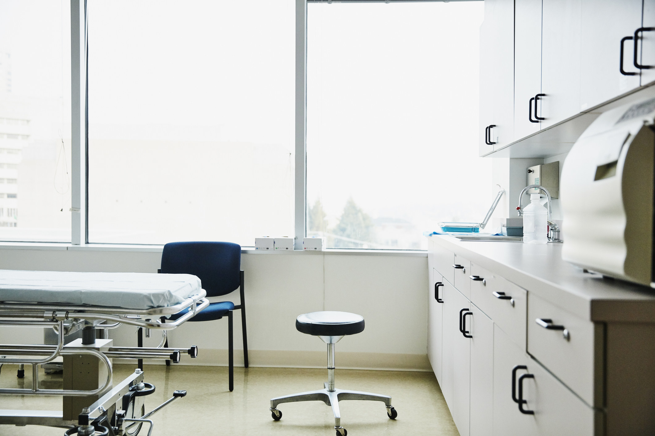 A bright and empty doctor&#x27;s patient room with a bed, short swivel chair, and countertop full of supplies