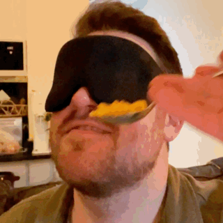 GIF of author&#x27;s partner feeding him mac &#x27;n&#x27; cheese while he wears an eye mask