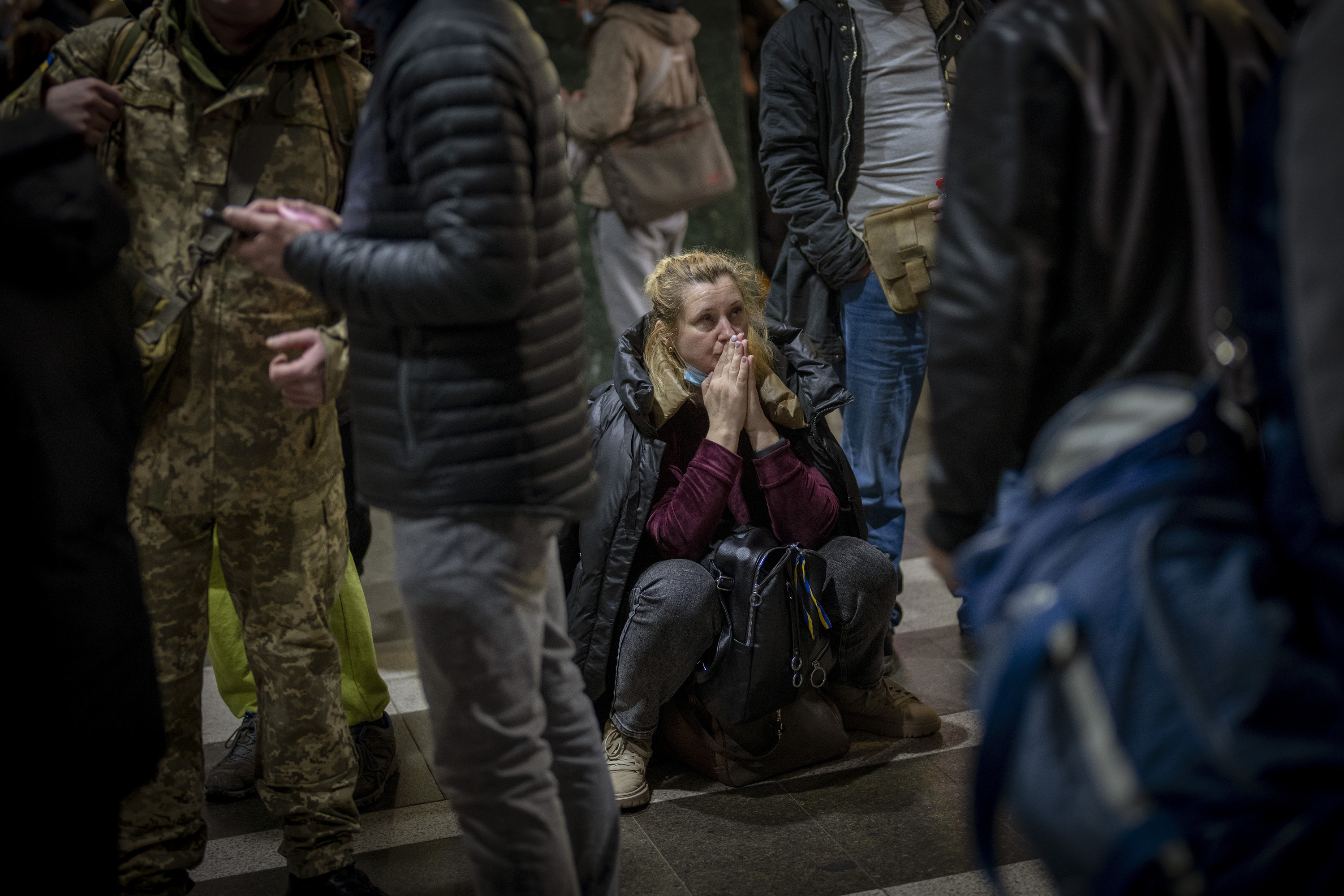 A woman crouches with hands folded, surrounded by standing people