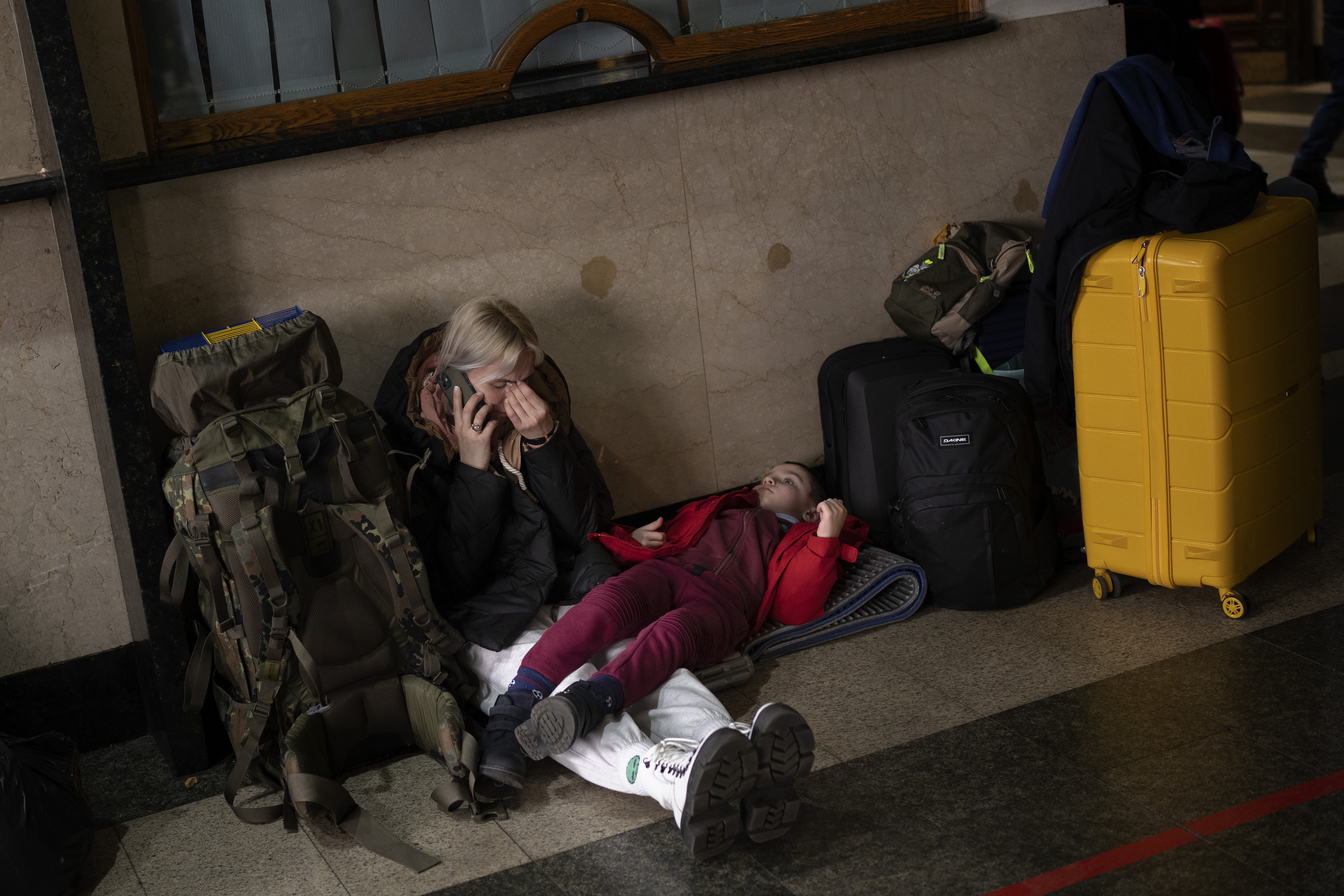 A woman pinches her forehead while on the phone while her daughter lies next to her, surrounded by luggage on the floor of train station