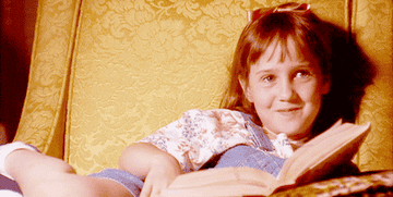 Young Matilda sitting in a gold armchair in dungarees, readings and laughing.