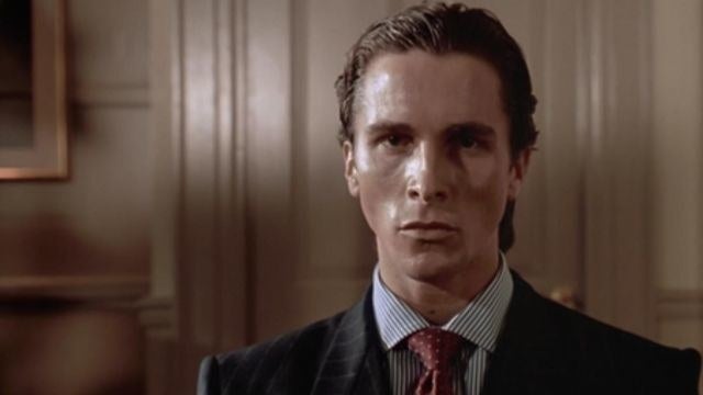 Person in a formal suit and tie, looking serious, standing in an office setting with wood paneling