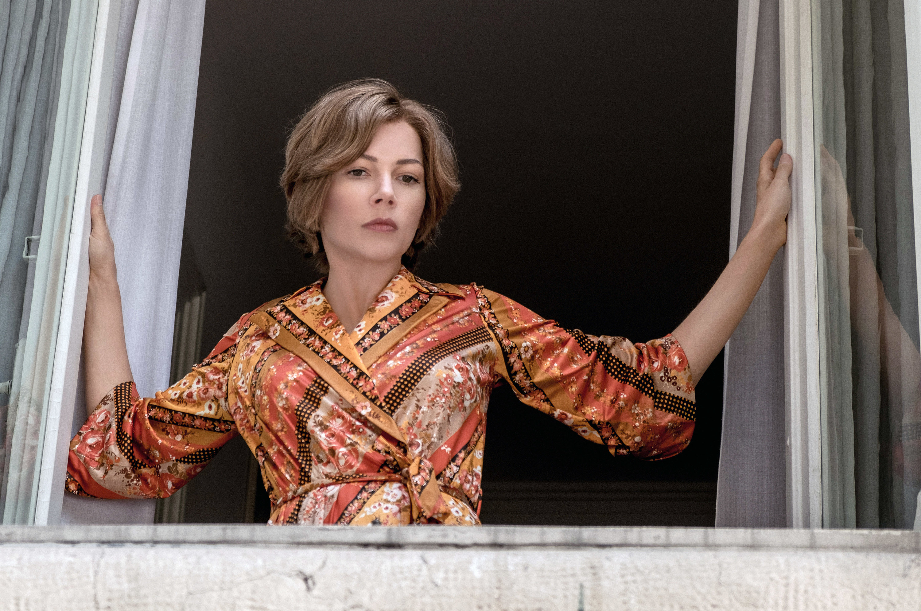 wearing an intricately patterned robe, Gail pushes open the curtains and looks down onto the street