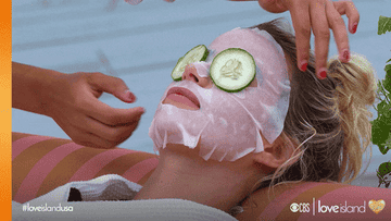 A person wearing a sheet mask with cucumbers on their eyes