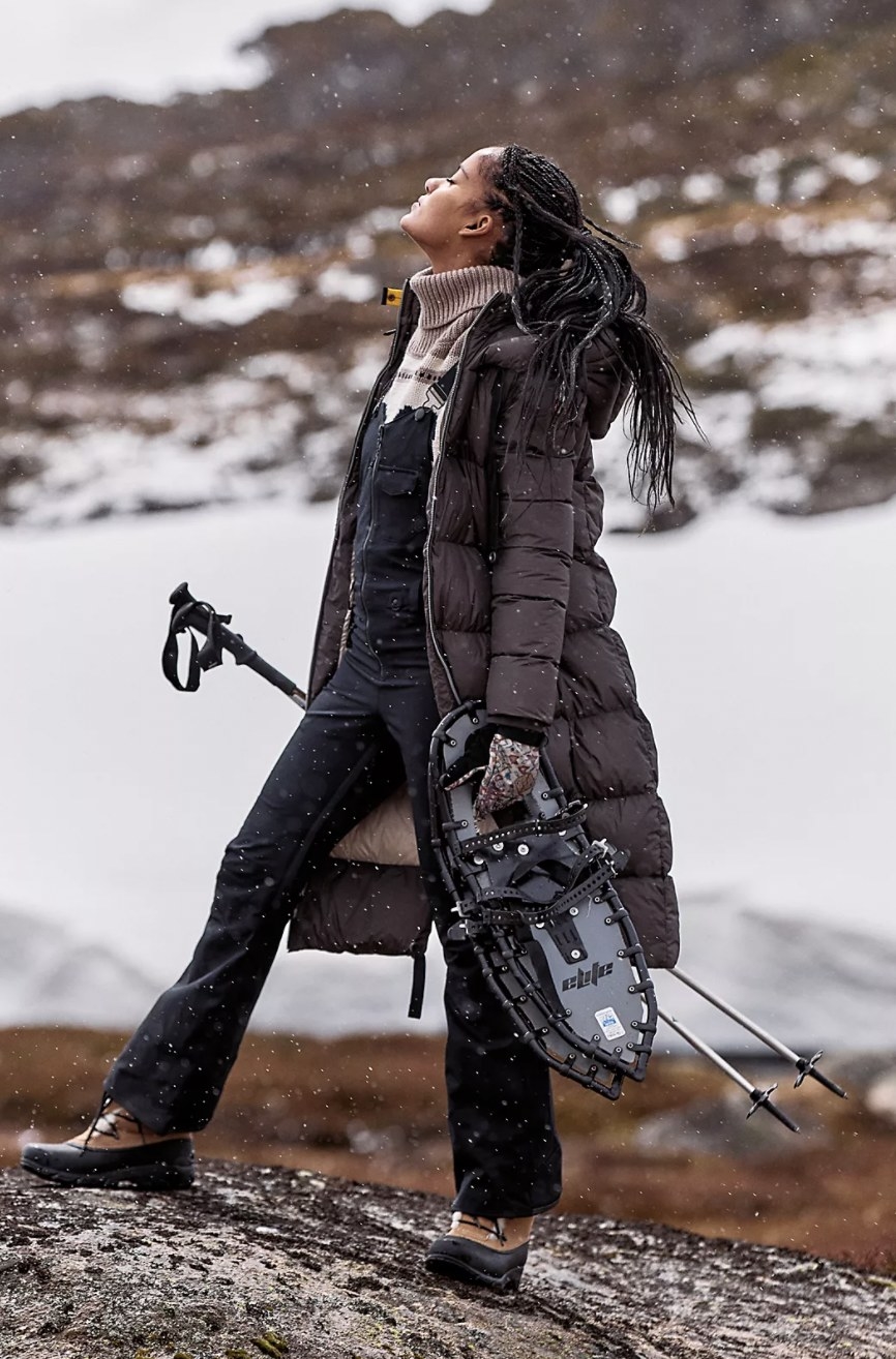 Model posing mid-hike in the black full-length coat carrying snowshoes and trekking poles