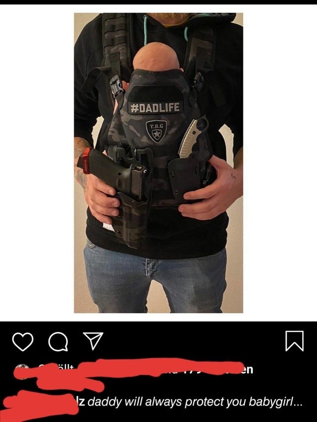 A photo of a dad holding a baby in a wearable baby carrier that also has holsters for a knife and a handgun