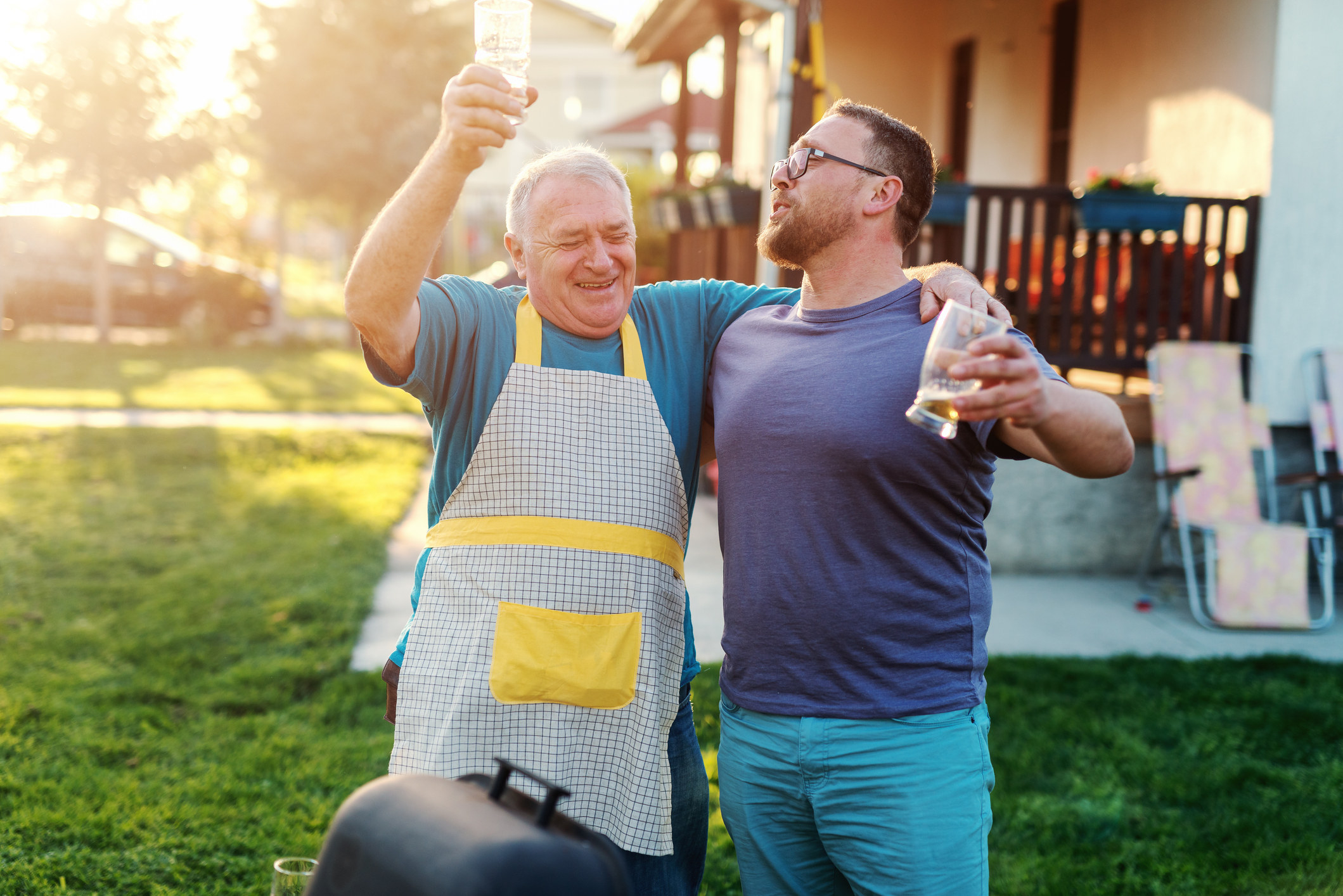 An older man with an apron hugs a younger man, with each of them holding a glass of beer