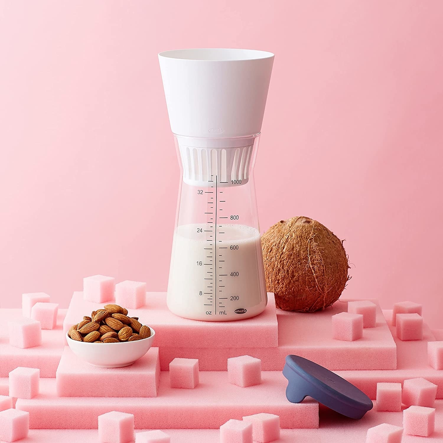 the hourglass-shaped nut milk maker next to fresh almonds and coconuts