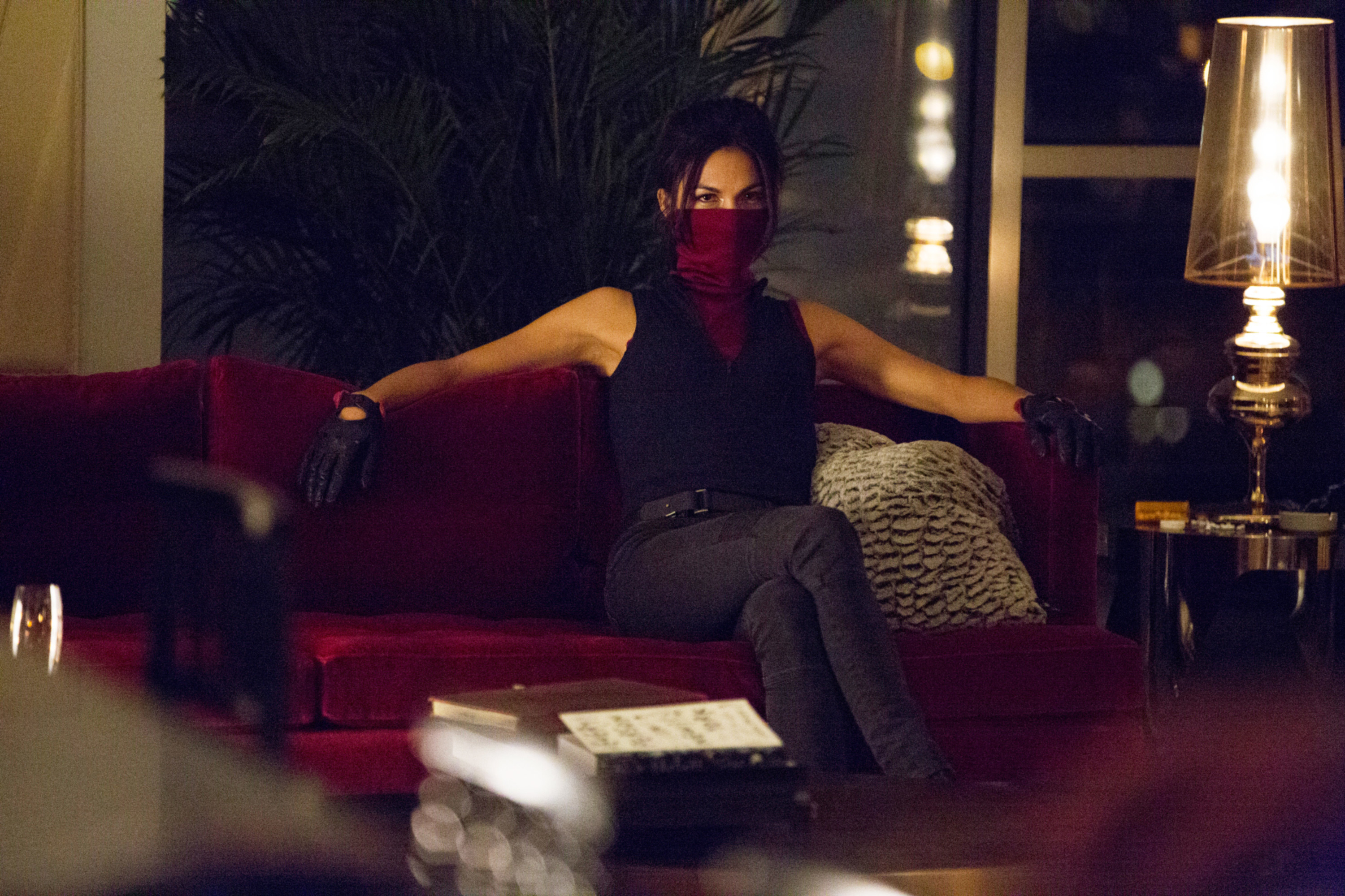 wearing a face covering, Elektra stretches her arm across the couch, waiting