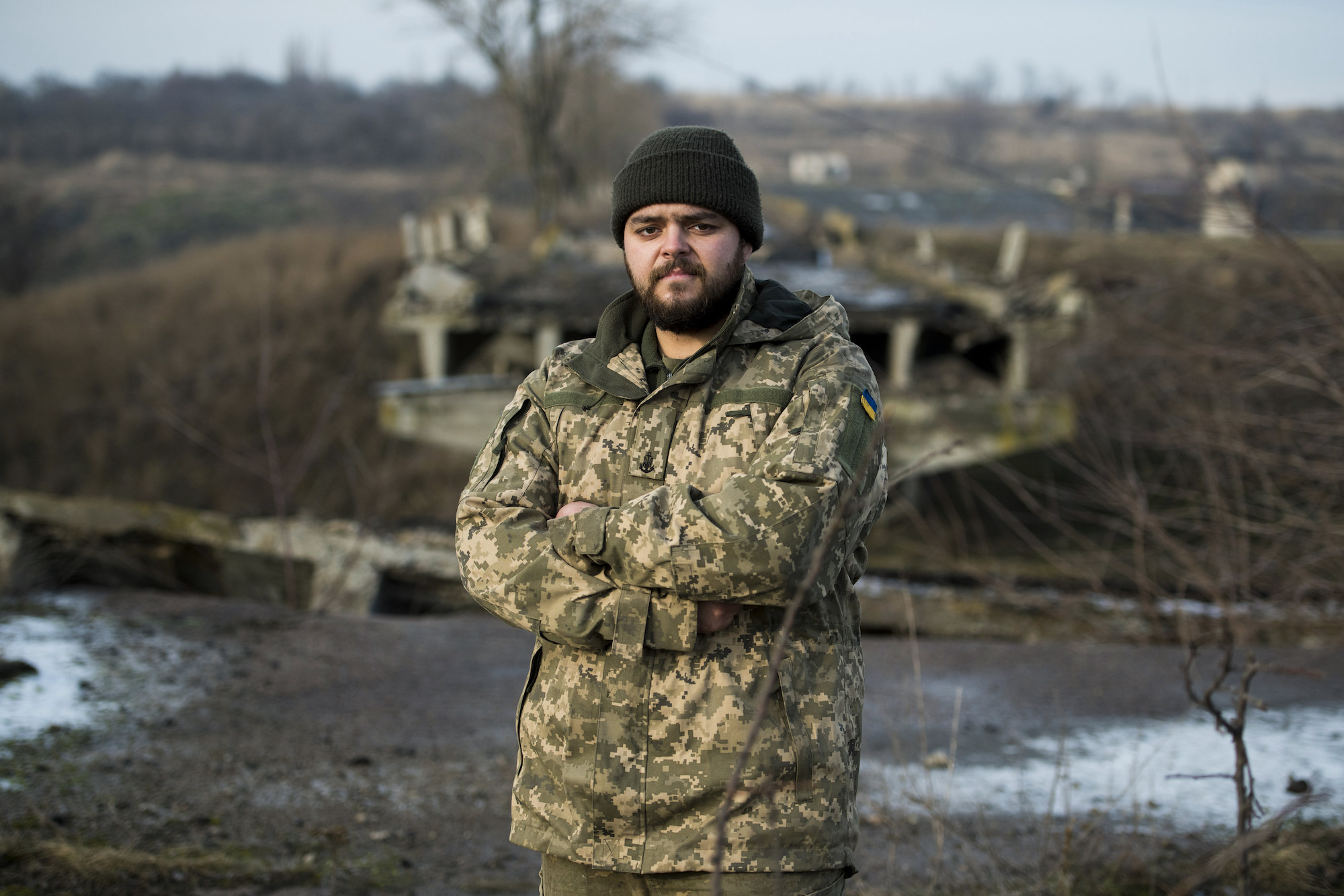 A man stands wearing fatigues and with his arms folded