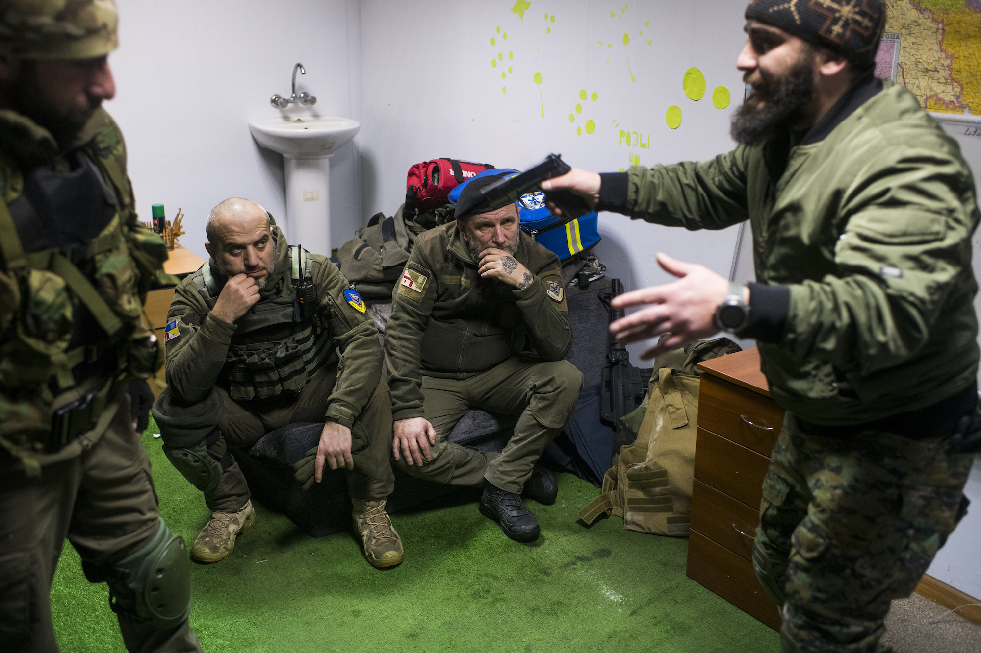 Two men sit as two men stand, one holding a weapon