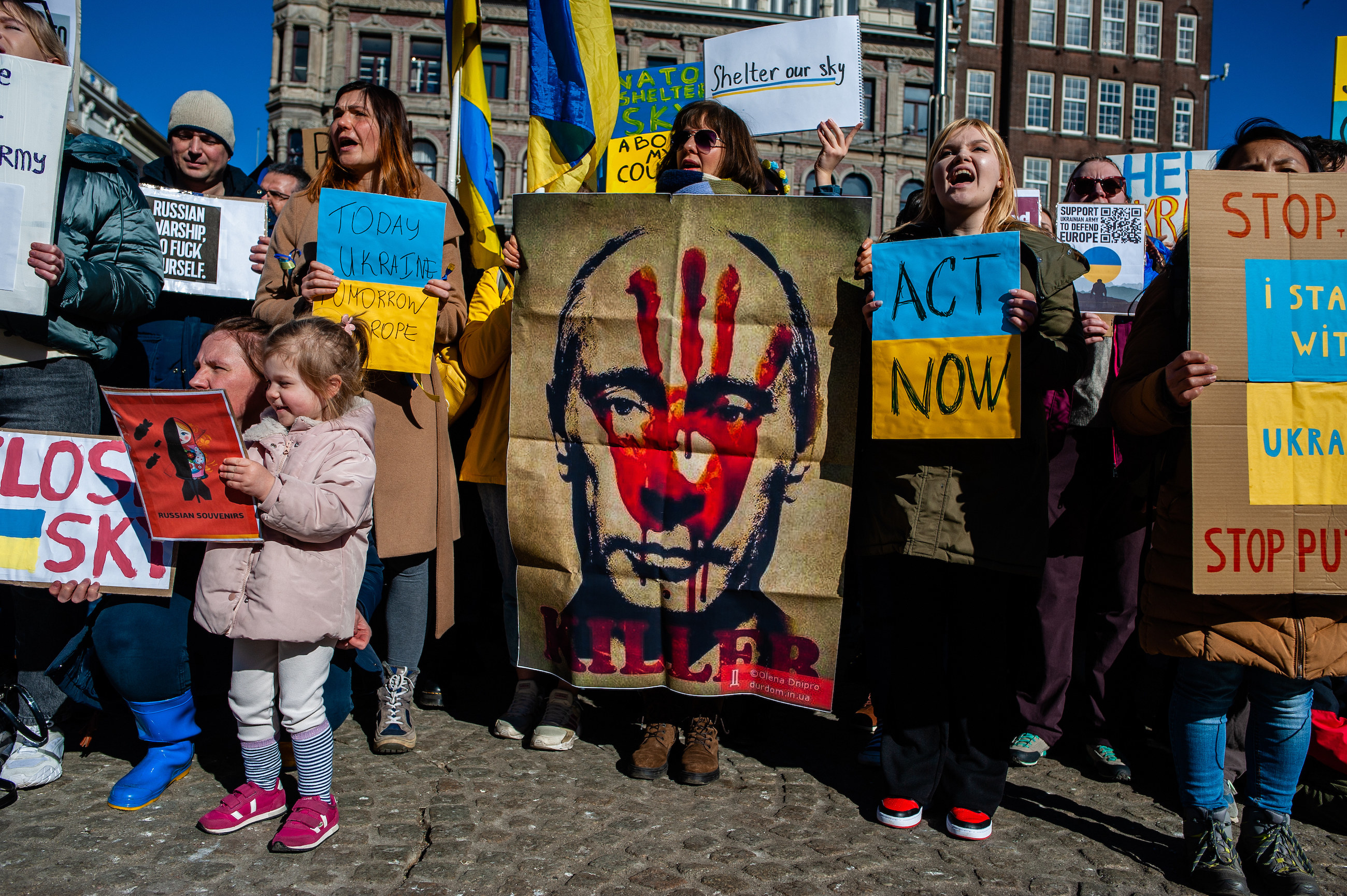A group of people, including children, holding signs, including one of Putin with a red handprint over his face