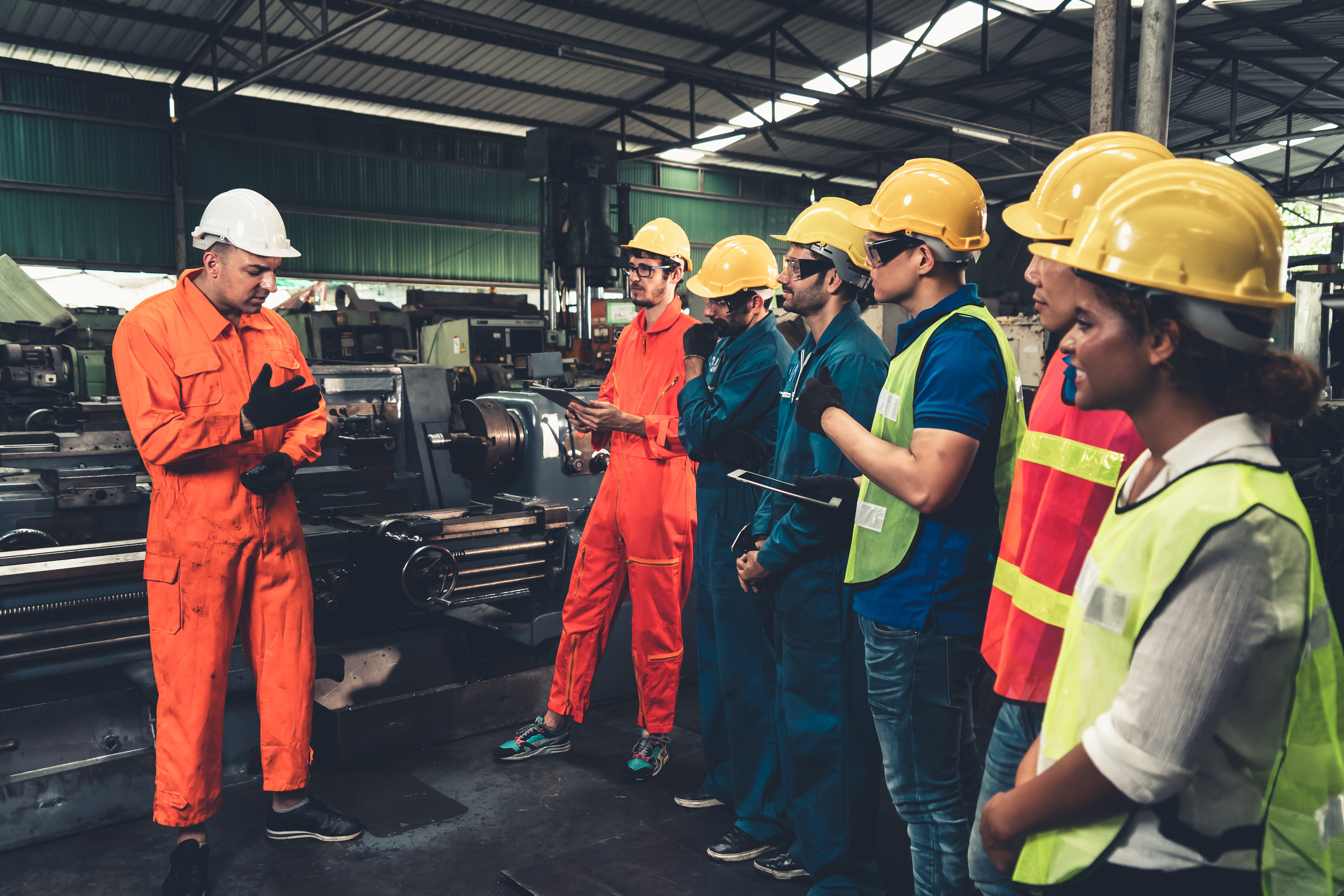 People wearing hard hats stand in a warehouse and listen to a person also wearing a hard hat