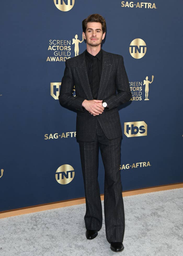 on the red carpet, Andrew wears a pinstriped suit and a classy watch, and he poses with his hands folded in front of him