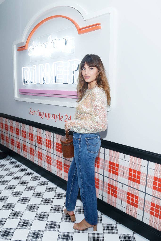 wearing jeans and a lacy blouse, Alyssa poses in front of a neon sign