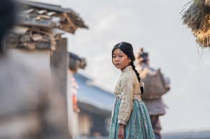 A young girl in a hanbok stands outside in Korea, looking forlorn