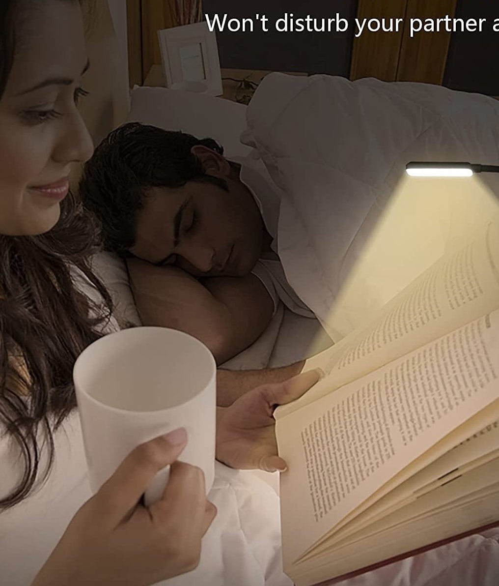 The lamp clipped to a book while a person reads in the dark next to their sleeping partner