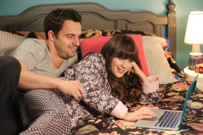 Jess and Nick spooning as they lie on a bed and look at a laptop