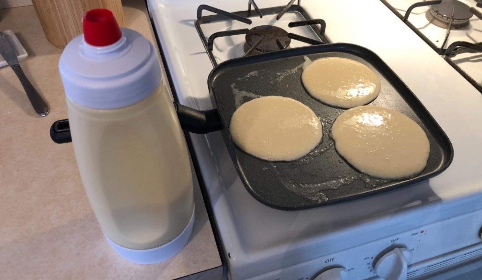 a reviewer photo of the dispenser filled with batter next to a griddle pan with pancakes cooking on it