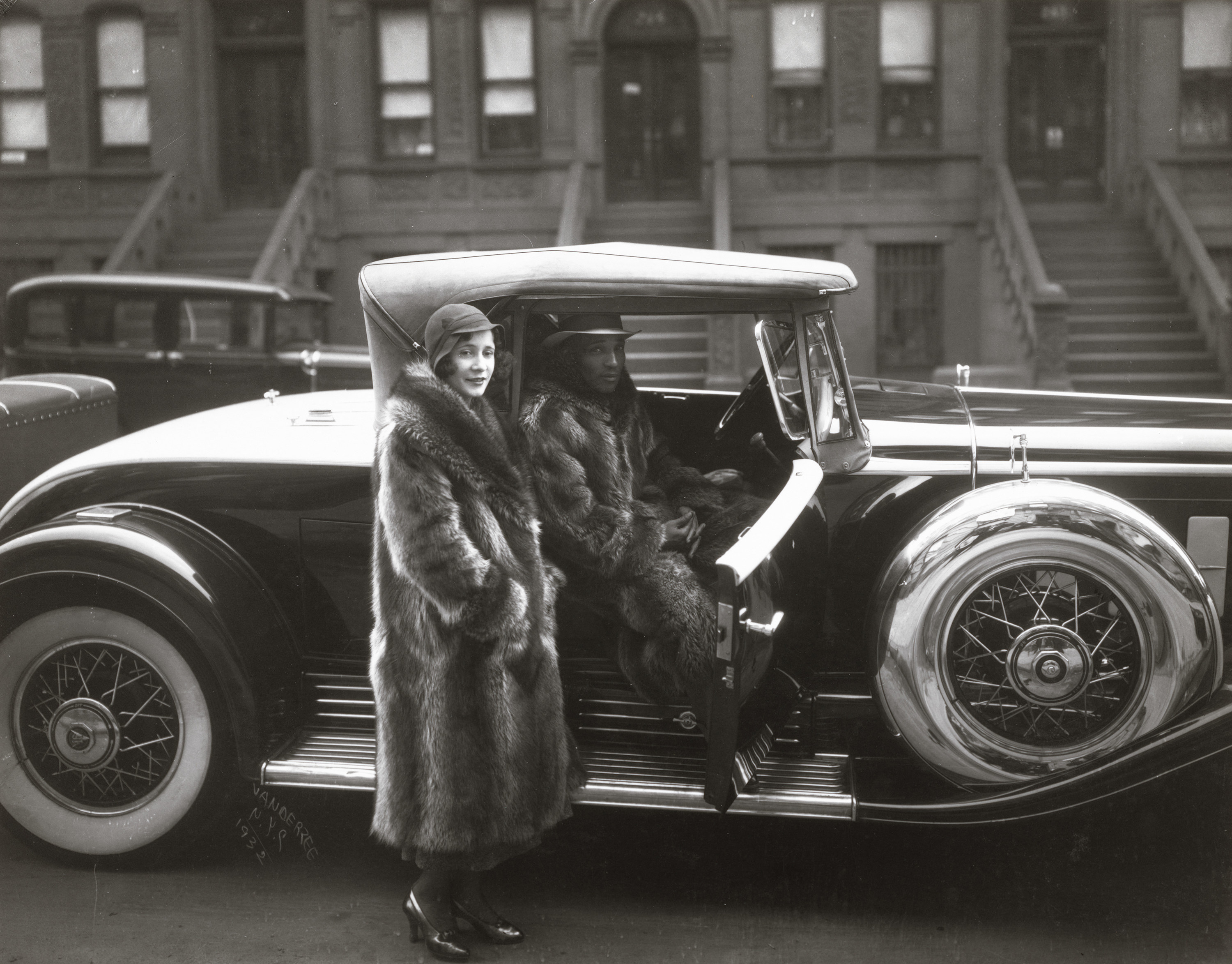 A black-and-white photograph of a couple in Harlem in 1932, with the man sitting in a car and the woman standing in a fur coat in front of it