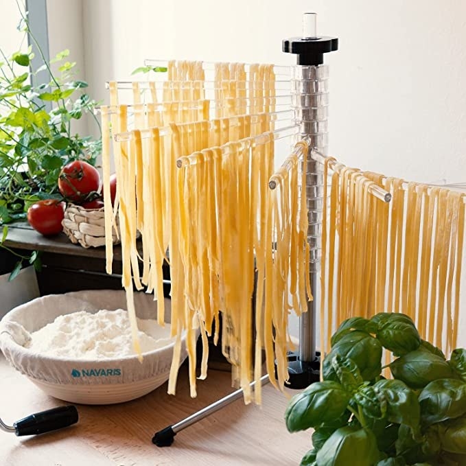 Fresh pasta noodles resting on the different levels of the rack next to a bowl of flour, and a pot of basil