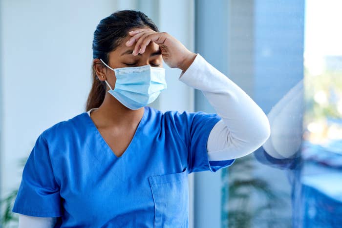 A nurse wearing a face mask and holding their head as they lean against a glass wall
