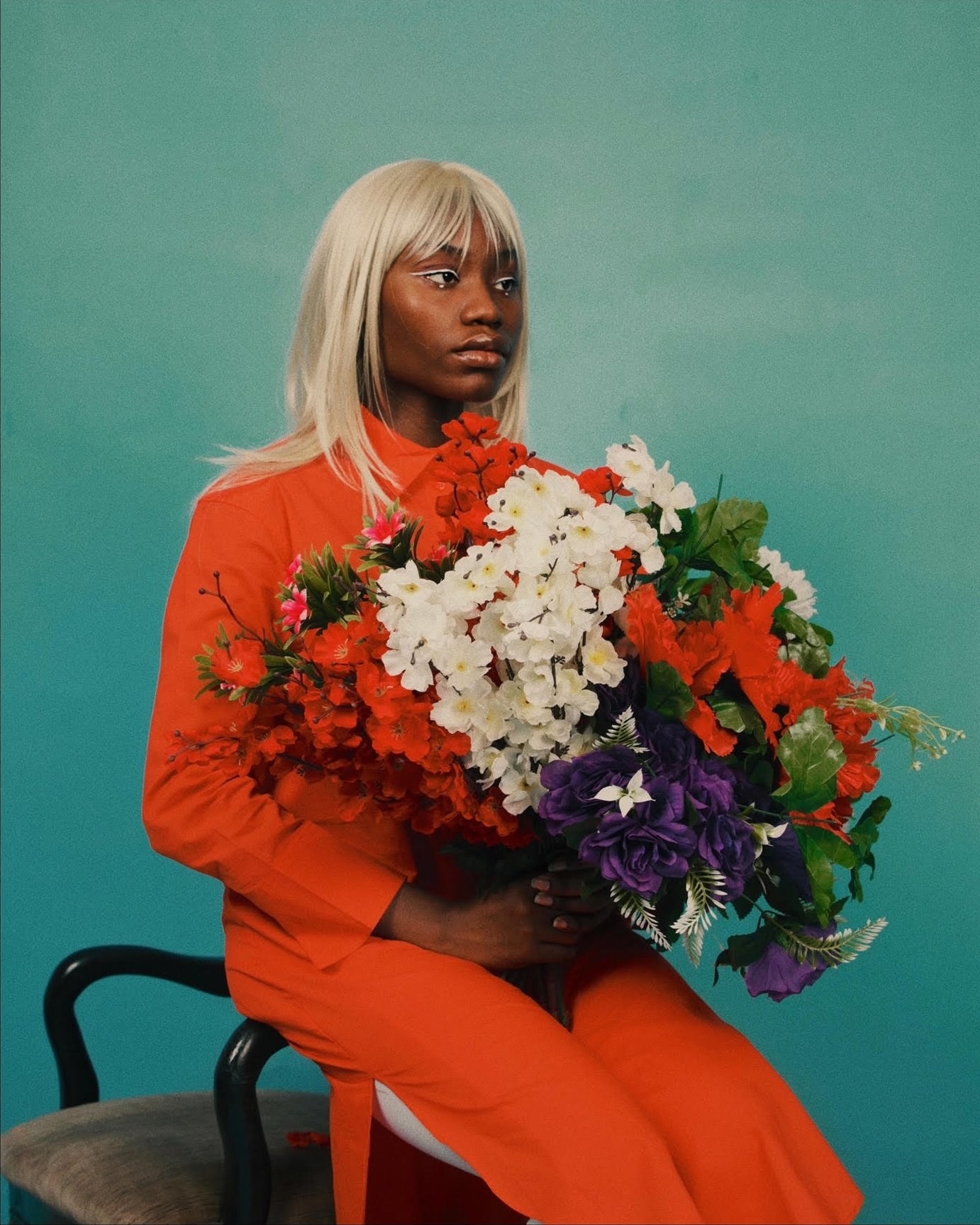 A person with blonde hair and vibrant eye makeup sitting on the arm of a chair and holding a large bouquet of flowers