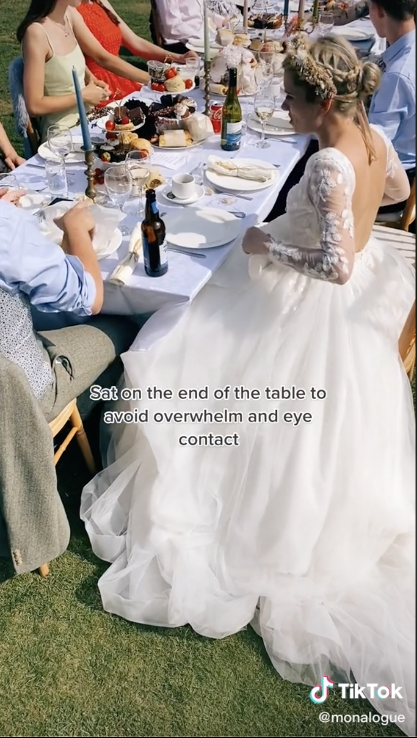 Screengrabs of a TikTok by user monalogue where a wedding party sits at a an outdoor table and Mona sits in a wedding dress at the end of the table, with a caption stating that she does so to avoid overwhelming and eye contact