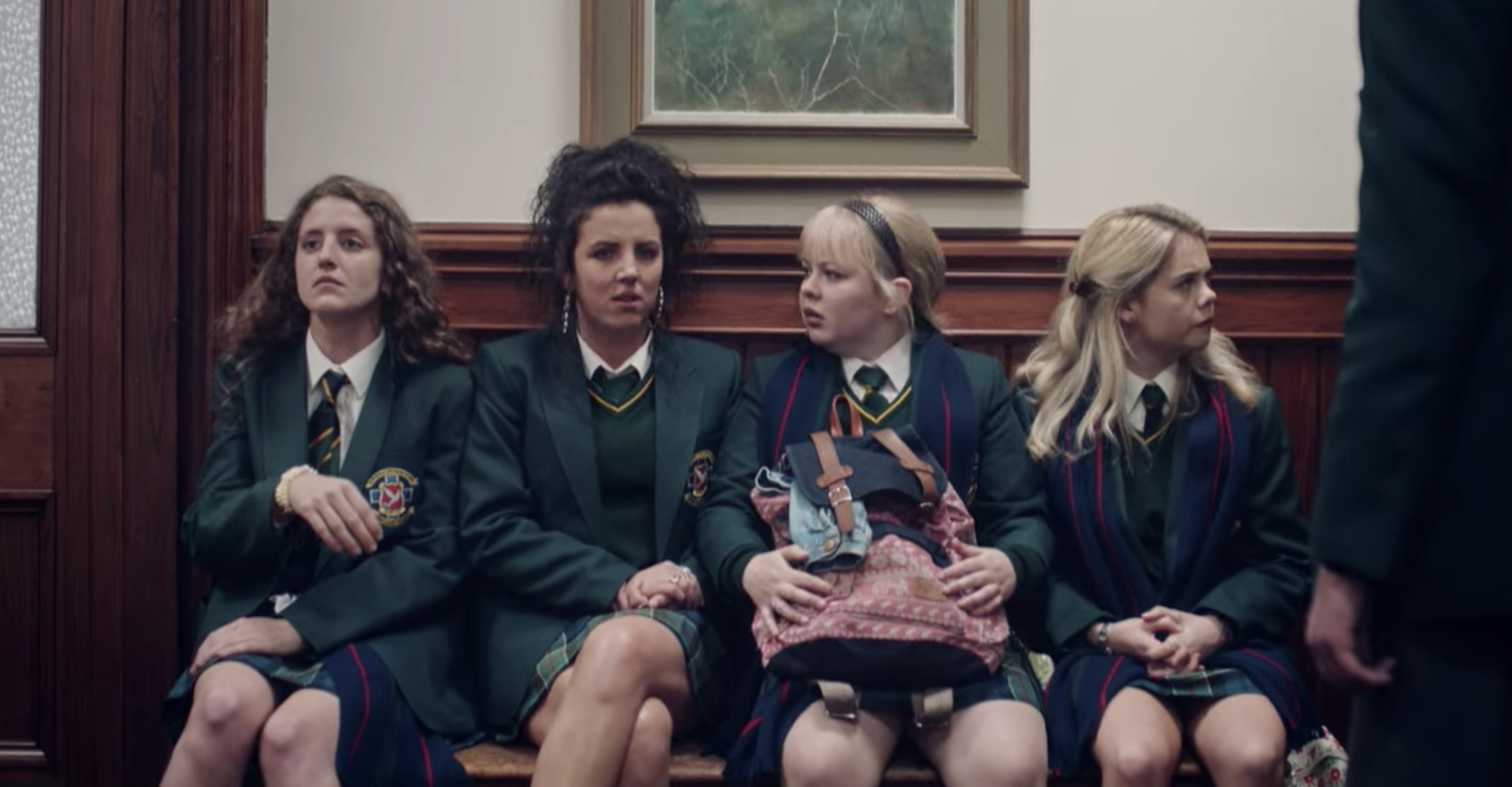 Nicola and three other girls dressed in school uniform and sitting on a bench in a scene from Derry Girls