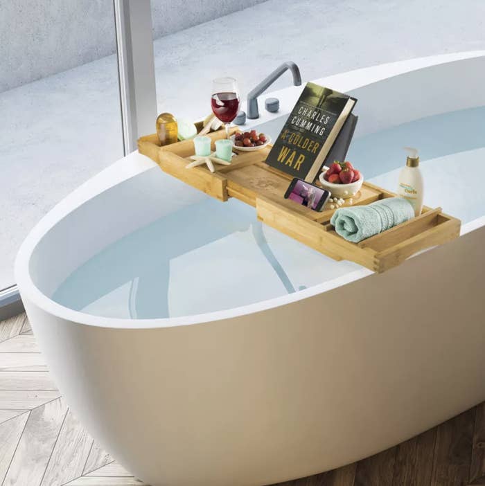the bath tray with a book, candles, a glass of wine, a phone and a towel on it over a full bathtub