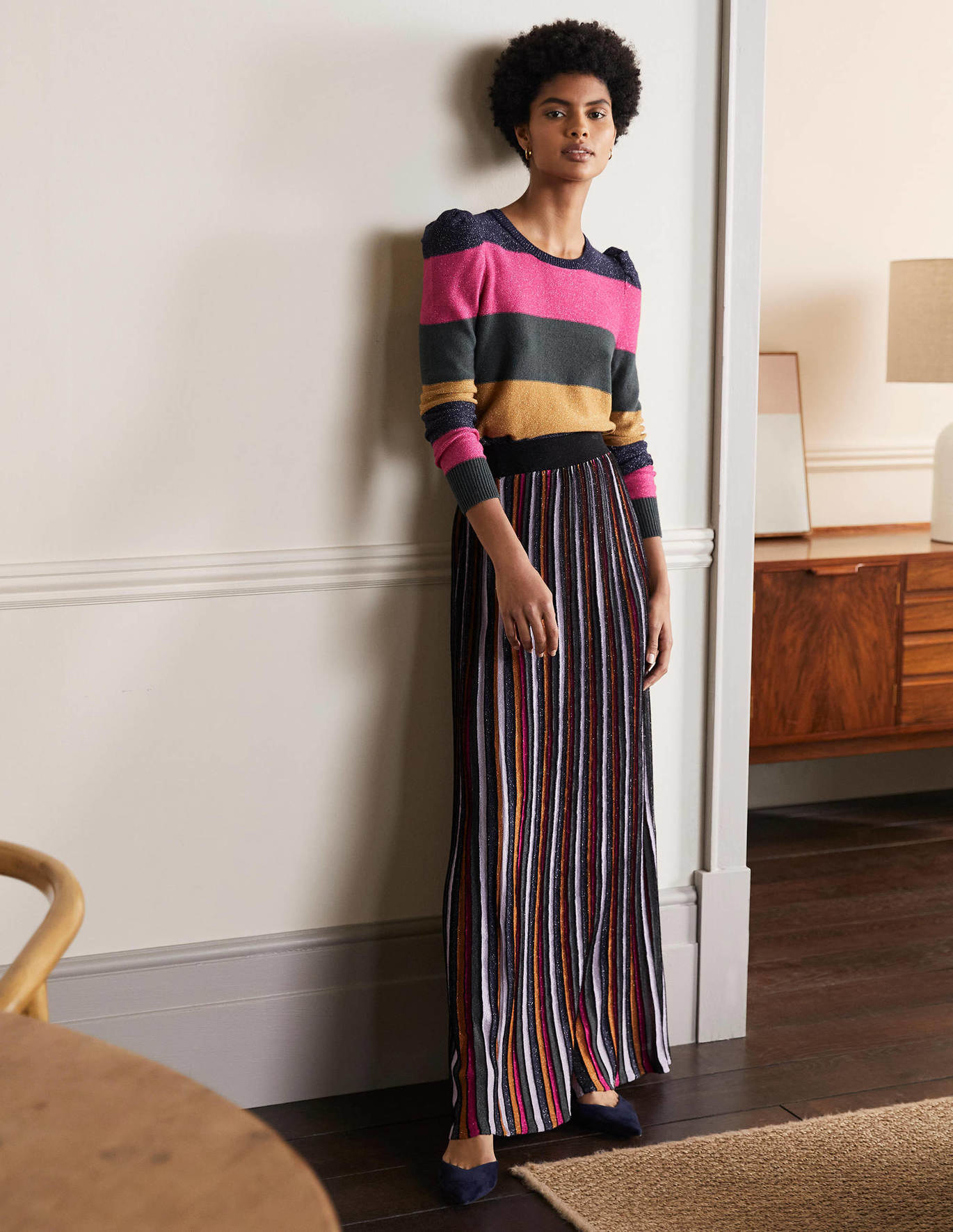 model wearing striped multicolor skirt, leaning against the wall in a house