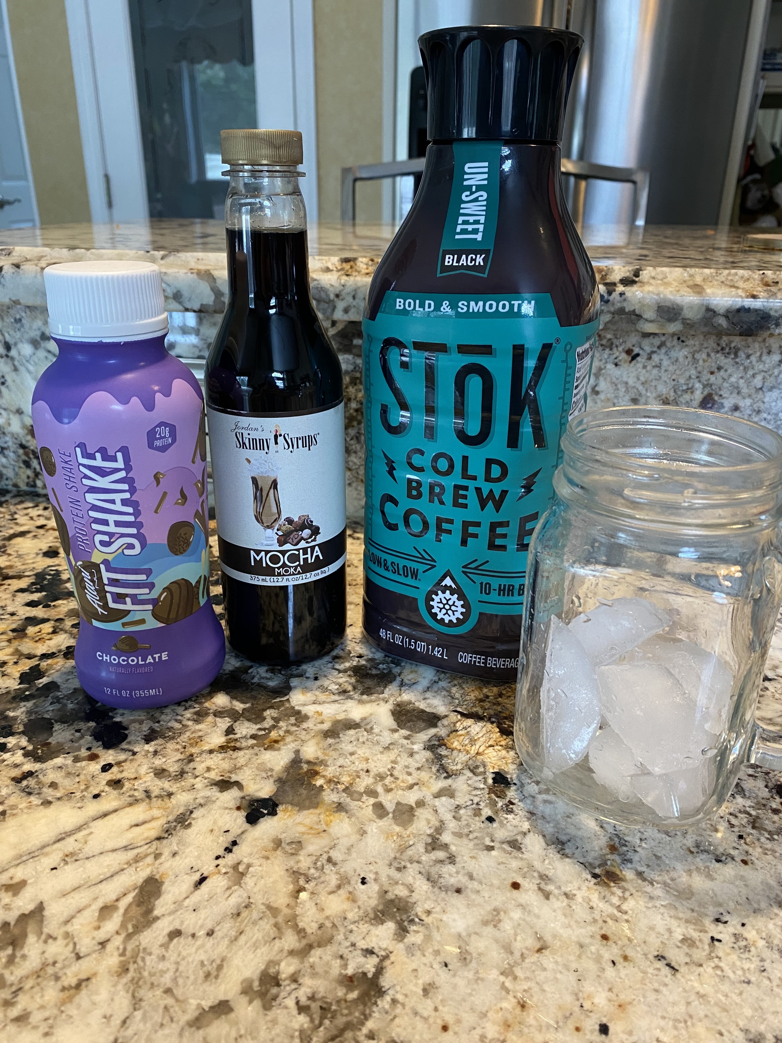 A lineup of a cold brew bottle, mocha coffee syrup, a chocolate protein shake, and a glass of ice