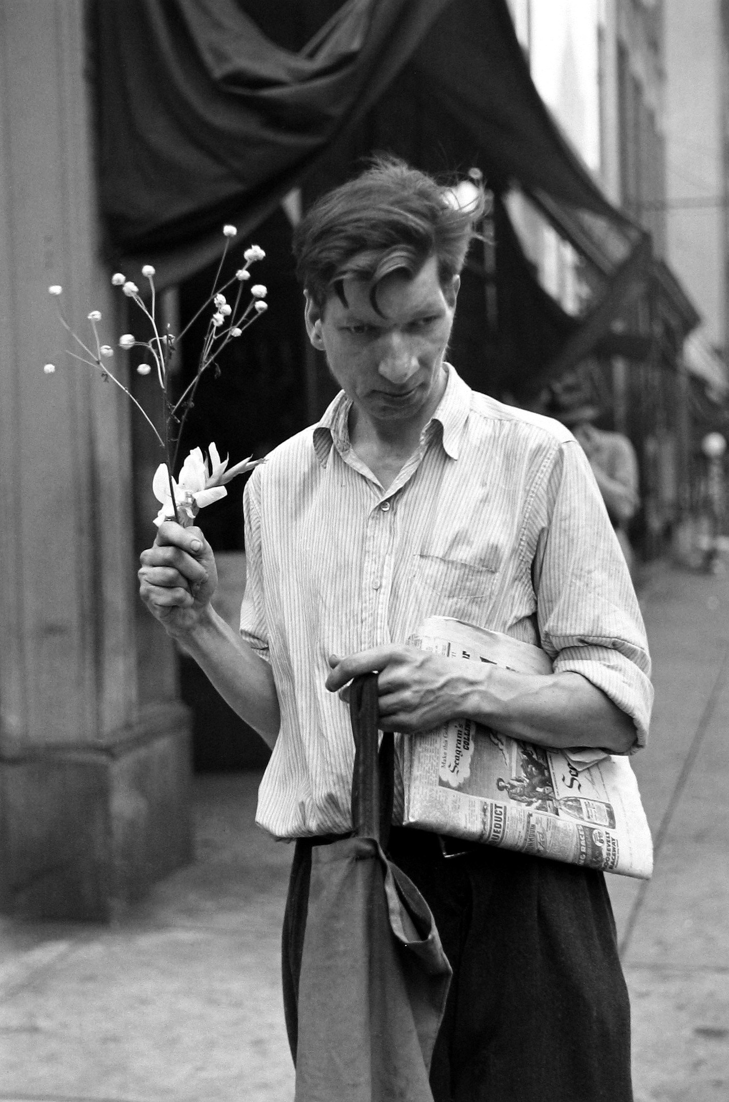 a man holds flowers, a newspaper and a tote bag and looks away from the camera