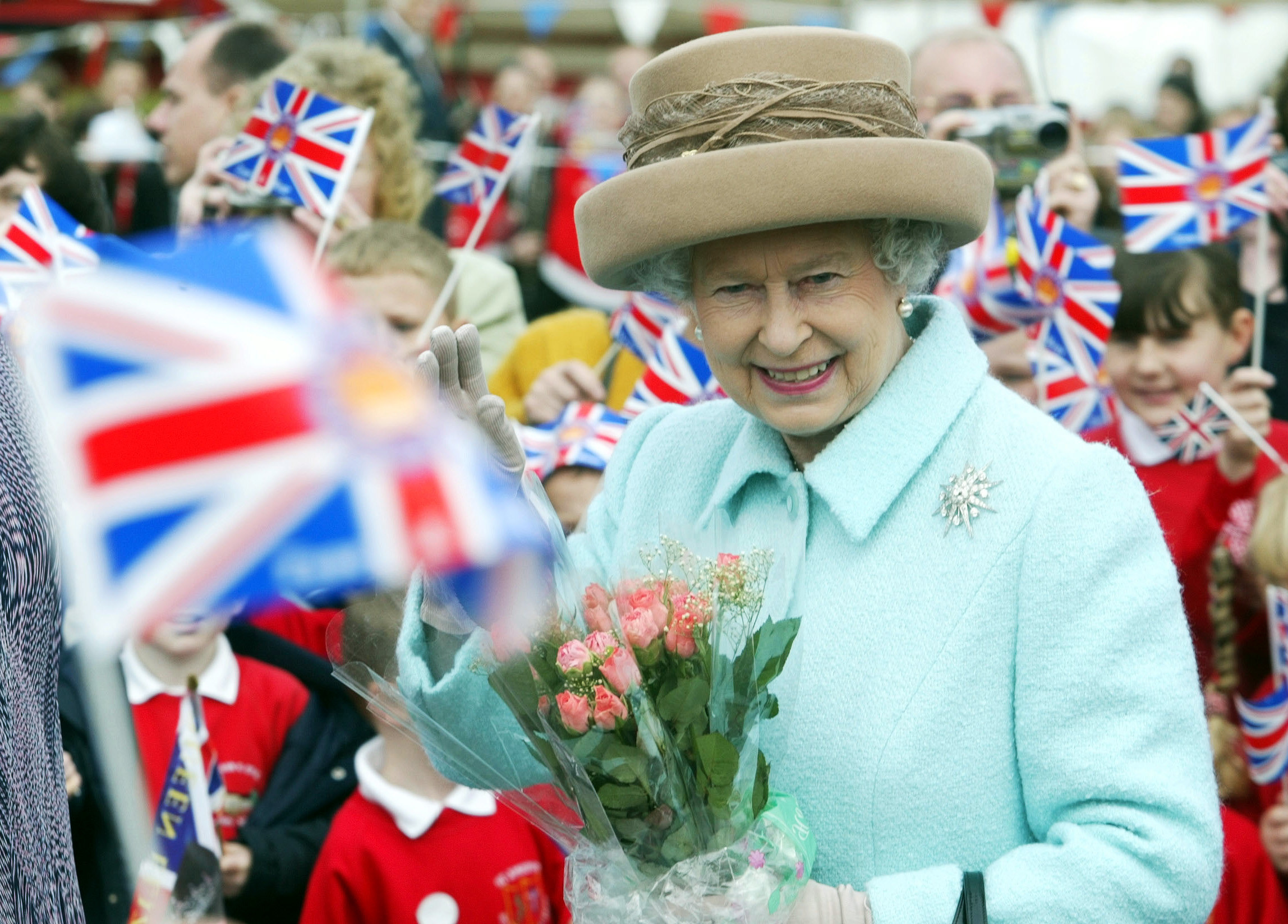 Queen Elizabeth II holding flowers among a crowd of people waving 
