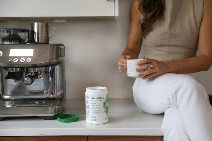 A photo of the lower half of a woman sitting on a kitchen counter holding a mug next to a coffee machine and a bottle of Organika.