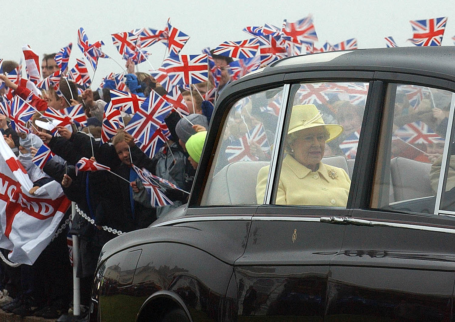 The queen in a car with a ton of people waving union jack flags surrounding her