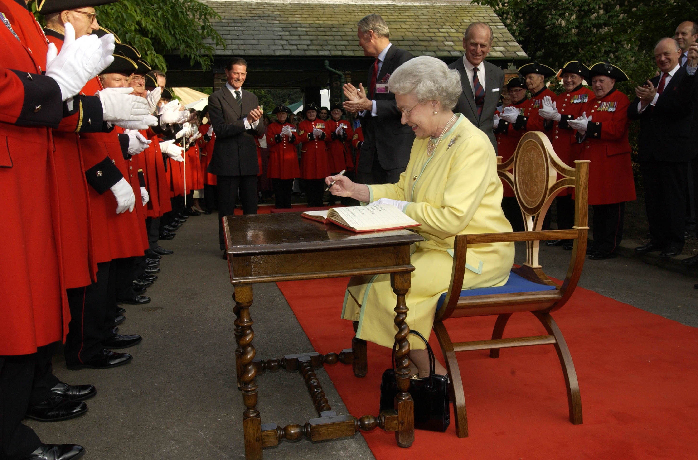 The queen sitting in a chair on a red carpet with people wearing red everywhere