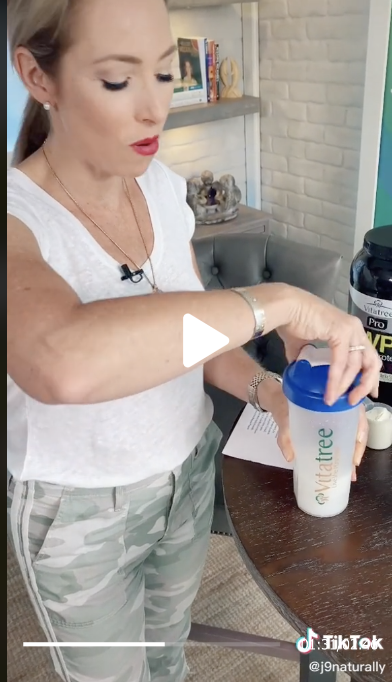 A woman in a white top and green camo pants holding a shaker bottle on a table
